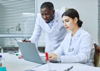Two researchers, a women seated, typing on a laptop computer and a man looking over her shoulder at the screen. They are both dressed in white lab coats and what appears to be a desk in white laboratory environment.