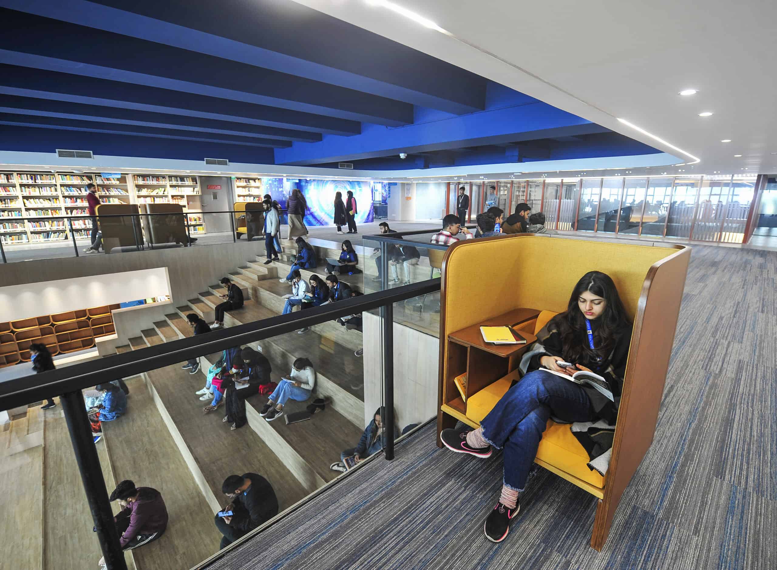 BRAC University Library. A student is seen studying in a yellow study booth, behind her is a large foyer room with books on the shelves and a wide staircase, on which other students are seated.